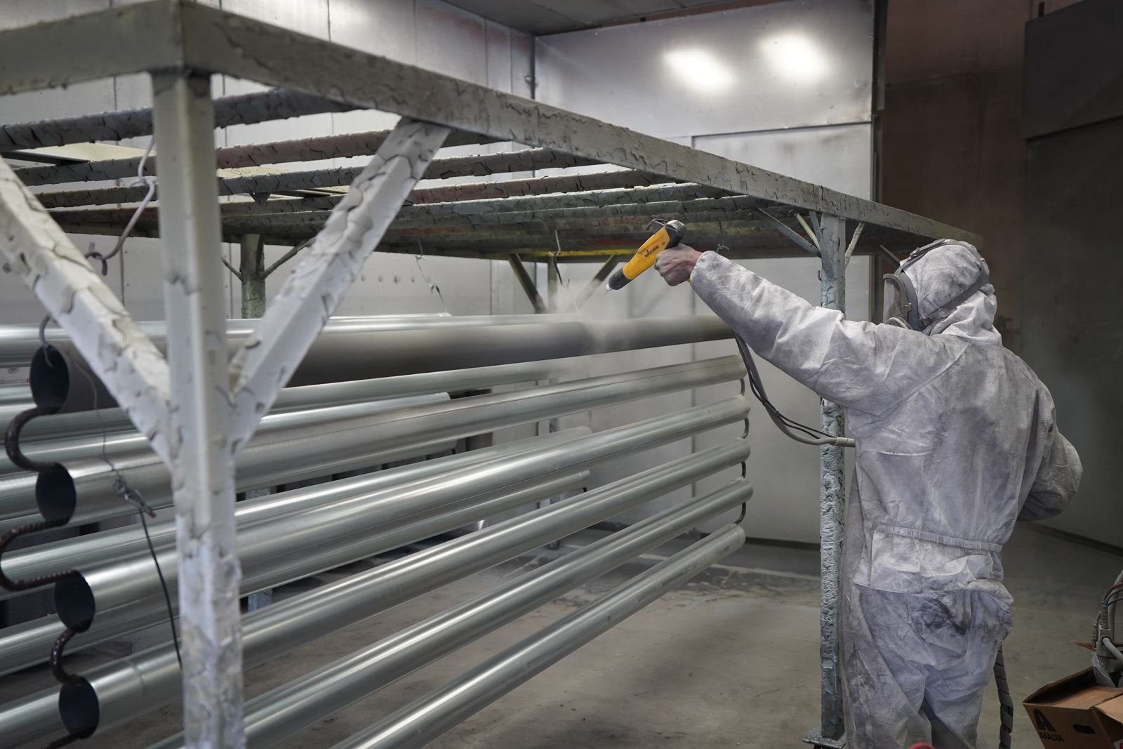 A technician in protective gear applying powder coating to metal components on a rack